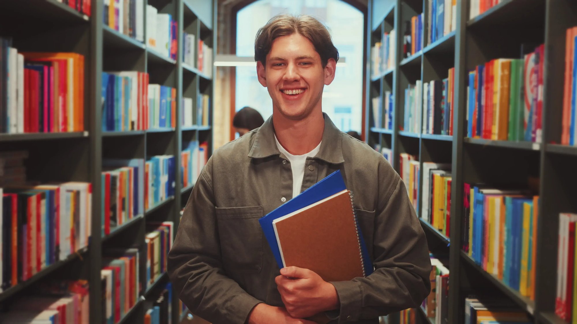 zoom-in-young-man-in-gray-jacket-smiling-and-holding-notebooks-in-library-aisle-lookin-SBI-351524122